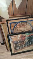 Framed pictures stacked against a wall including a Miller Genuine Draft advertising poster in a black frame, a colorful vintage artwork in a gold frame, and another piece partially visible.