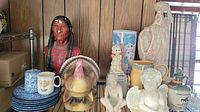 Wide view of entire pottery lot including Native American bust, turkey figurine, plates, mugs, and various ceramic pieces on wooden surface.