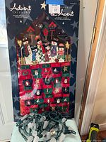 Photo of fabric wall hanging advent calendar shaped like a house with 24 numbered red and green pockets featuring snowman and Christmas motifs. Includes attached string lights.