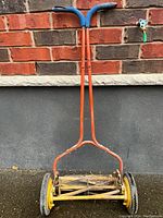 Full front view of the antique push lawn mower showing the orange frame and blue foam handle grips against brick wall background.