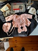 Overview of crocheted pink baby clothes (jacket and bonnet), white bonnet, Little Bo Peep baby cutlery set in ceramic holder, and Peg-Pérego rain cover packaging.