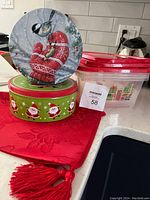 Photo showing two round Christmas-themed tins, a rectangular plastic container with a red lid and Christmas gift print, and a red fabric table runner with tassels, all arranged on a kitchen countertop.