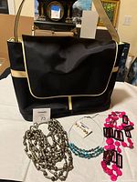 Black fabric computer bag with gold trim, displayed with various jewelry including metallic chain necklace, turquoise beads, pink earrings, and stud earrings.