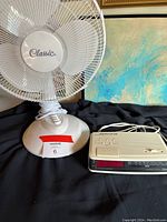 Photo of a white Classic brand small oscillating fan and a beige digital clock radio with control buttons, both placed on a black cloth surface in front of a wall with a painting.