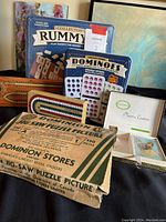Photo showing vintage Dominion jig-saw puzzle in original box with Dominion stores branding, collector's Rummy and Dominoes games in tin boxes, and vintage plastic playing cards in a white box