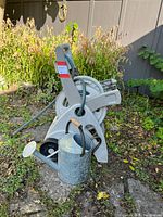 Photo showing a grey hose caddy with a 50-foot green garden hose coiled around it, placed outdoors on soil and leaves with greenery in the background. A galvanized metal watering can with wooden handle is positioned in front.