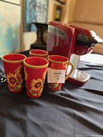 Red Keurig coffee machine seated on table alongside four red ceramic sunflower mugs, each with a gold rim.