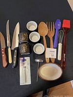 Photo of assorted kitchen utensils including knives, ramekins, wooden utensils, whisk, basting brush, measuring spoons, napkin rings and ceramic dish on black surface.