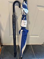 Photo showing two large umbrellas standing side by side, one black with curved handle wrapped in plastic, the other blue and white with tag.