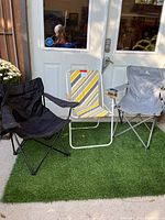 Three folding lawn chairs positioned outdoors on artificial grass in front of a white door: one black fabric chair, one vintage striped yellow and white metal-frame chair, and one gray Outbound branded folding chair.