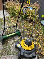 Both lawn mowers situated outdoors on a paved area with leafy surroundings: a green Lee Valley push reel mower and a Black & Decker electric lawn mower with yellow extension cord on top.