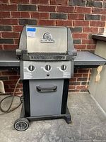 Front view of Broil King Monarch BBQ with three burner control knobs and lid with logo. Two side shelves and wheels visible.