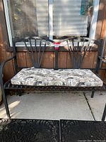 Black metal outdoor bench with floral cushion on the seat, positioned in front of a window on concrete floor.