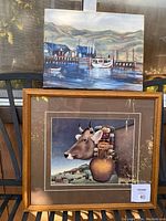 Photo showing two artworks: an original oil painting depicting a harbor scene with boats and hills, and a framed print of a cow with a decorative arrangement on its head.