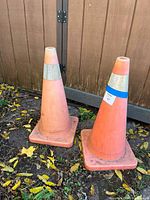 Two large orange traffic pylons with reflective bands standing outside against a wooden fence on dirt ground with leaves.
