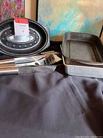 Photo showing oval speckled black roasting pan with lid, rectangular dark roasting pan, and BBQ tongs, spatula, and fork.