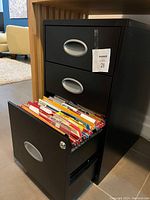 Front view of small black metal filing cabinet with all drawers closed except bottom drawer open showing hanging folders inside.