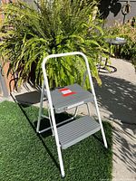 Photo of a foldable step stool with two grey ribbed steps and white metal frame, standing outdoors on a patio with plants and a table in the background.