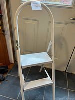 White two-step metal stool with a rounded handle, visible paint stains on the legs and steps.