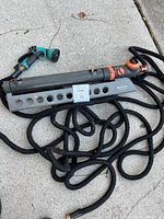 Image shows a large oscillating sprinkler lying on pavement connected to a long black stretchy hose and a black and teal handheld spray nozzle.