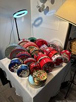 24 assorted Christmas-themed cookie tins of various sizes and colors, arranged on a white cloth-covered table with lamps and chair in background.