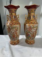 Pair of Asian ceramic vases with decorative handles and scenic figures against intricate patterned background on a white surface with shadow cast.