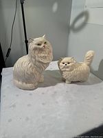 Two white ceramic cat figurines displayed on white cloth, one larger cat with green eyes and one smaller cat with black eyes, chip visible on large cat ear