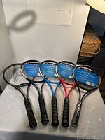 Photo showing five tennis rackets laid out flat, clearly showing grips and frames in black, blue, and red colors.