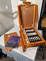 Closed portable wooden art easel standing on a table, case open revealing paint tubes and brushes inside, with a pack of palette paper and wooden palette board beside it.