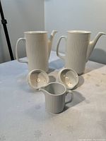 Two white ceramic coffee pots with vertical ribbed texture and lids, alongside one matching creamer, displayed on a white tablecloth.