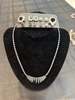 Close-up image of a pewter necklace with rectangular beads and fan-shaped pendant displayed on black stand with card showing six pairs of earrings including flower designs with clear and light blue stones.