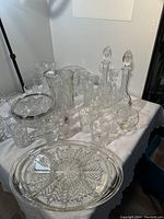 Full lot of assorted crystal and glass bowls, trays, decanters, pitchers and glasses arranged on a table covered with a white cloth, displaying the pattern variety and quantity.