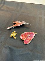 Three brooches on black fabric: coral hummingbird with rhinestones, yellow bee with rhinestones, red-pink leaf with gold edge