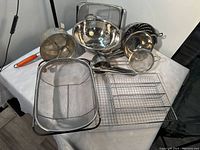 Photo showing the assortment of metal kitchen tools including a large mesh strainer, colanders, wire racks, and a food mill arranged on a table.