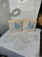 Display of four clear glass snack sets showing oval trays and matching cups with original Homestead boxes in background.