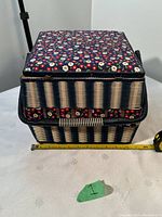 Front view of woven sewing basket showing floral fabric-covered lid and woven basket sides along with measuring tape for scale.