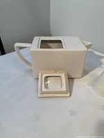 Side view of the square white ceramic teapot without its lid, showing the open square opening and handle design.