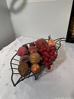 Wire basket filled with assorted frosted artificial fruit seen from a side angle against a plain wall and white fabric surface.
