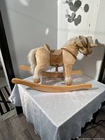 Full view of the small child's rocking horse showing plush body, white mane and tail, leather straps, and wooden rockers on white table with sunlight.