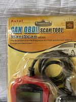 Front view of scan tool inside original packaging showing red handheld device with screen and attached cable.