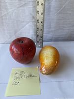 Two marble fruit figurines, one red apple and one yellow pear, shown with ruler for size reference.