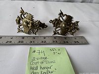 Two vintage brass coat of arms wall hangers or door knockers placed side by side next to a ruler showing measurements.