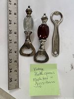 Photo shows the three vintage bottle openers placed next to a ruler for size reference, approximately 5 inches long each. One opener has a metal handle with maple leaf engraving, and the two others have decorative glass handles, one clear and one maroon, with ornate finials on top.