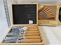 Full view of the 7-piece carving tool set in an open natural wood box with foam lining, blades wrapped in plastic, and a ruler beside for scale.