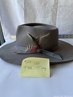 Front view of the grey felt hat with feather on the side and handwritten note showing measurements 9" tall 15" long.