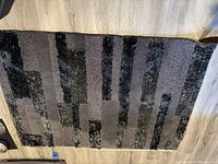 Black rectangular area rug with vertical blocks and strips of different texture and height, laid on a wooden floor.