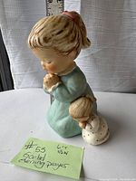 Photo of Goebel Hummel figurine with measuring tape showing height approximately 6 inches, little girl kneeling in prayer holding a doll, light blue dress.