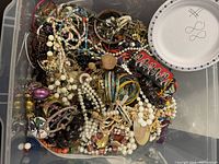 Top-down view of large plastic container filled with assorted crafting jewelry including beaded strands, cords, metal chains, and various components.