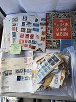 Photo showing a pile of various stamp collection pages, loose stamps inside a large plastic bag, some envelope pieces with stamps, and a red vintage 'Majestic Stamp Album'.