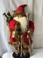 Front view of large Santa figure showing red coat with brown faux fur trim, white beard, and detailed face. Holding wooden stick staff with bell and a rustic lantern. Burlap sack with greenery visible on back.